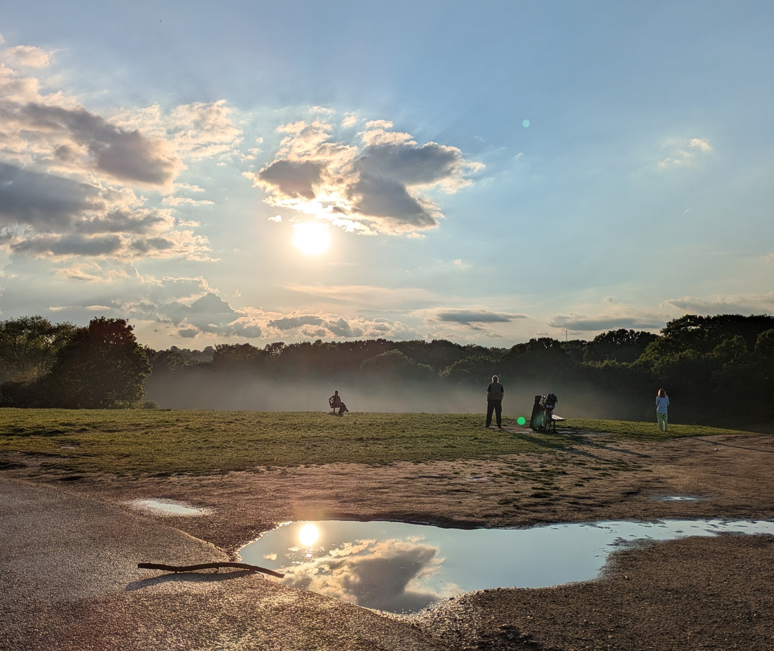 Top of a hill with lots of ground mist, setting sun, people looking into the sunset framed by the sun in the rising mist, and a puddle with a reflection of the sun behind a handful of clouds.