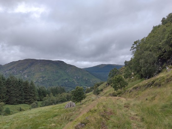 Photo of a hilly landscape with a grassy path, trees on the right, green hills in the distance and a cloudy sky
There is also a weirdly split stone further down the path, as if struck by lighting some time ago, I'm not sure what happened to it!