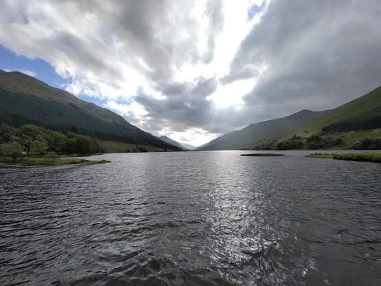 Photo of the loch taken from the shore with small ripples in the blue-grey water,green hills on both sides in the distance and still a cloudy sky but with the sun coming through in places